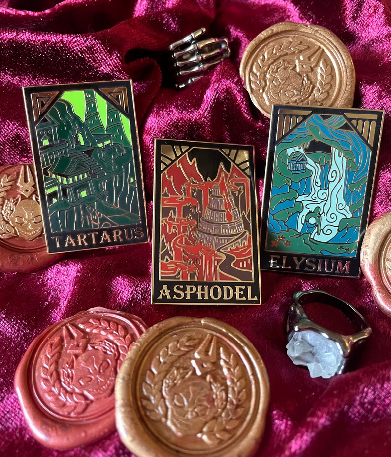 May include: Enamel pins with gold trim and the words "TARTARUS", "ASPHODEL", and "ELYSIUM". The pins are on a burgundy velvet surface, along with gold wax seals and a skeleton hand ring.