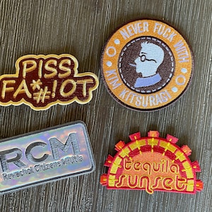 Può includere: Quattro toppe ricamate con vari disegni e testo. Una toppa dice "PISS FA*#!OT" in marrone e beige. Un'altra dice "NEVER FUCK WITH KIM KITSURAGI" con una silhouette. Una terza toppa recita "RCM Revachol Citizens Militia". L'ultima toppa dice "tequila sunset".