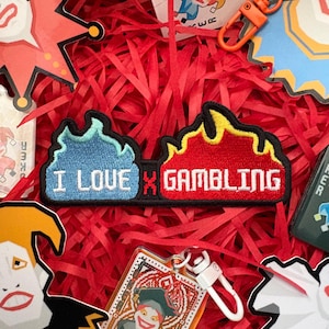 Puede incluir: Primer plano de accesorios con temática de juego. Destaca un parche con el texto "I LOVE GAMBLING" en diseño de llama. Otros elementos incluyen naipes, un llavero y recortes decorativos, todo sobre un fondo de papel rojo triturado.