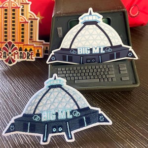 May include: Two stickers featuring a dome-shaped building with the text "BIG MT" in blue and white. One sticker is placed on a vintage computer, and the other is on a wooden surface. A third sticker of a building is also visible.
