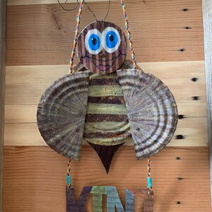 May include: A whimsical bee made from recycled materials hangs from a string with the word "KIND" written on a wooden sign below. The bee has a brown body, blue eyes, and yellow and brown striped wings.