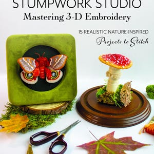 May include: A book cover titled "STUMPWORK STUDIO Mastering 3-D Embroidery" features a 3D embroidered moth and mushroom. The book is about 15 realistic nature-inspired projects. Scissors, leaves, and embroidery thread are also visible.