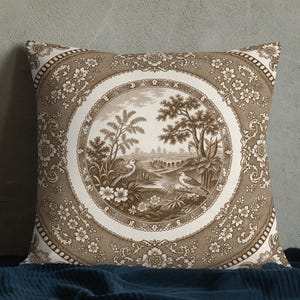 May include: A square throw pillow featuring a brown and white toile pattern. The centre displays a circular medallion with a detailed landscape scene of trees, birds, and foliage, surrounded by a decorative floral border on a brown background.