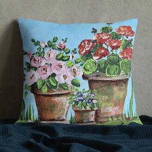 May include: A throw pillow with a blue background featuring a painting of three potted plants with pink, red, and purple flowers. The plants are in brown pots with green leaves.