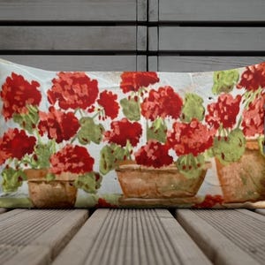 May include: Rectangular cushion featuring a watercolour design of red geraniums in brown pots. The flowers have green leaves and are set against a white background. The cushion is a home decor item.