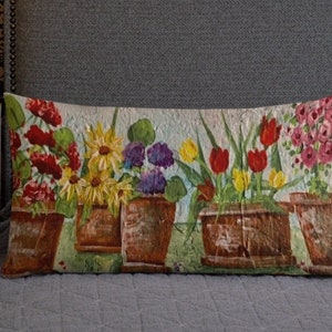 May include: A rectangular pillow with a floral design. The pillow features a row of five flower pots with different flowers in each pot. The flowers are painted in shades of red, yellow, purple, and green. The background of the pillow is a light beige color.