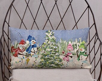 May include: A decorative throw pillow with a winter scene featuring five snowmen decorating a Christmas tree. The snowmen are wearing colorful hats and scarves. The background is a blue sky with falling snow.