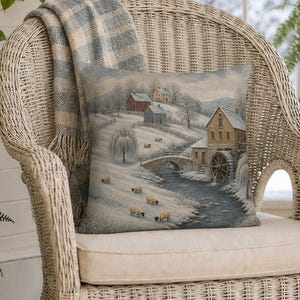 May include: A square throw pillow featuring a woven winter landscape scene with a water mill, a stone bridge, and sheep grazing on a snow-covered hillside. The pillow is placed on a white wicker armchair draped with a blue and white plaid throw blanket.