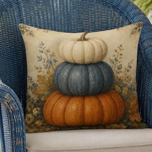 May include: A square throw pillow featuring a printed illustration of three stacked pumpkins in cream, blue, and orange. The background displays a botanical pattern of dried leaves and flowers in muted earth tones, set against a light beige fabric.