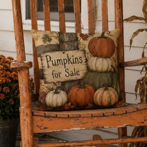 Puede incluir: Una mecedora de madera naranja desgastada con un cojín cuadrado que presenta un gráfico de varias calabazas y el texto Pumpkins for Sale. La silla está situada en un porche con un farol, un crisantemo en maceta y tallos de maíz secos.