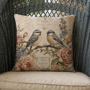 May include: A square throw pillow featuring a beige background with two blue and brown birds perched on a branch, surrounded by pink and blue floral illustrations, with the text Paris and Jardin de Fleurs printed on the front surface.