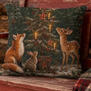 May include: A square throw pillow featuring a printed scene of a fox, a rabbit, and a fawn gathered around a decorated Christmas tree. The dark green background highlights the animals and the tree, which is adorned with glowing lights and ornaments.