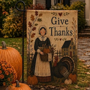 May include: A rectangular garden flag featuring the text Give Thanks, an illustration of a person in a bonnet and apron holding a basket of pumpkins, a turkey, and a house, all set against a cream background with autumn leaf accents.
