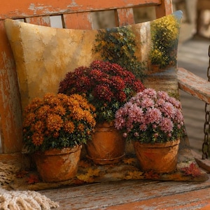May include: A square throw pillow featuring a printed scene of three terracotta pots filled with blooming chrysanthemums in shades of orange, deep red, and soft pink, set against a warm, textured background of a stone wall and greenery.