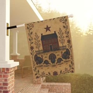 Puede incluir: Una bandera de jardín rectangular con fondo color canela, una casa roja, una estrella de color rojo oscuro y cuatro pavos en primer plano. El diseño está enmarcado por un borde de enredaderas marrones, hojas y pequeñas bayas rojas, colgado de un poste blanco.