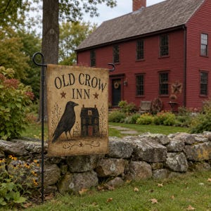 May include: A rectangular garden flag featuring a beige background with the text "OLD CROW INN" in black lettering. The design includes a black crow illustration, a small house graphic, and two brown stars, all suspended on a black metal stand.