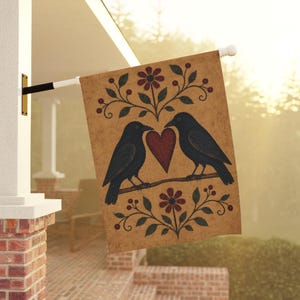 Puede incluir: Una bandera de jardín rectangular con fondo color canela que muestra dos cuervos negros posados en una rama frente a frente. Entre los cuervos hay un corazón rojo. Arriba y abajo de las aves hay diseños florales simétricos con flores rojas y hojas de color verde oscuro.