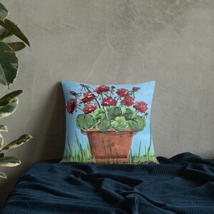 May include: A throw pillow with a painting of a potted plant with red flowers and green leaves on a blue background.