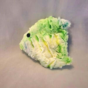 May include: A plush, abstract-shaped pillow in shades of green and white, with a black button detail. The soft, textured surface creates a fluffy appearance. The pillow is designed with a unique, artistic form.