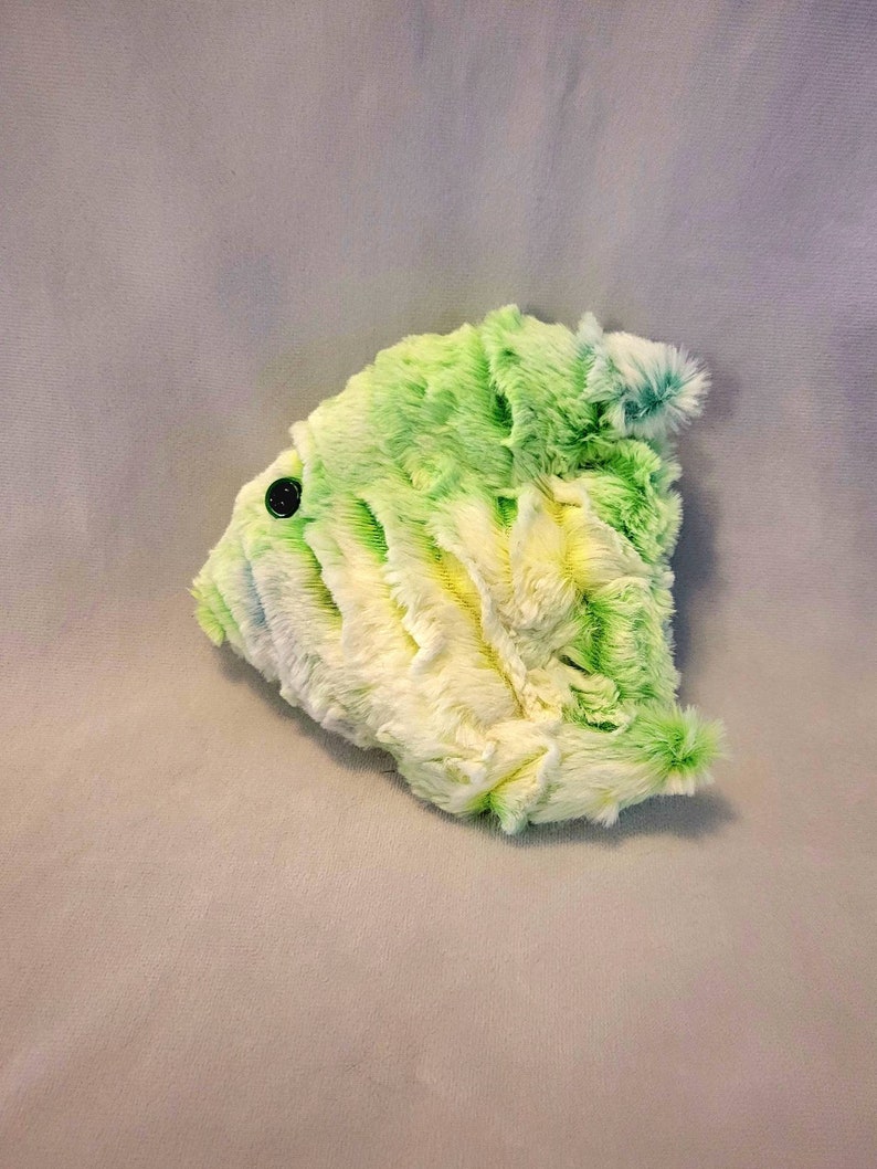 May include: A plush, abstract-shaped pillow in shades of green and white, with a black button detail. The soft, textured surface creates a fluffy appearance. The pillow is designed with a unique, artistic form.