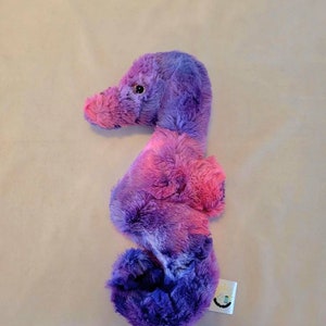 May include: A plush seahorse toy in shades of purple and pink. The stuffed animal has a curved body, a long snout, and a small, round eye. The seahorse is made of soft, fluffy material.