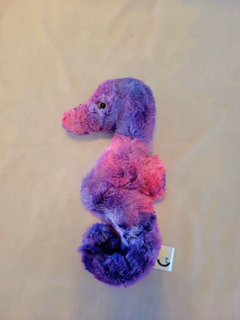 May include: A plush seahorse toy in shades of purple and pink. The stuffed animal has a curved body, a long snout, and a small, round eye. The seahorse is made of soft, fluffy material.