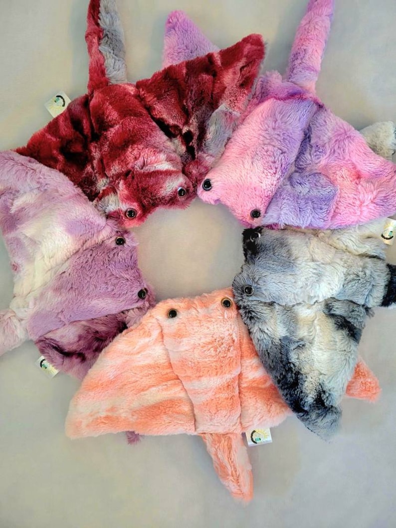 May include: A collection of plush stingray toys in various colors, including red, pink, purple, and gray. Each toy has button eyes and a tag. The toys are arranged in a circular pattern.