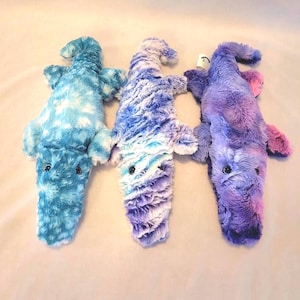 May include: Three plush alligator toys in shades of blue, purple, and pink. Each toy has a textured, furry appearance with small black eyes. The alligators are lying flat, showcasing their body shape and color variations.