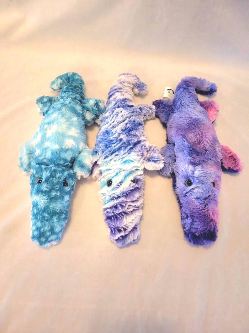 May include: Three plush alligator toys in shades of blue, purple, and pink. Each toy has a textured, furry appearance with small black eyes. The alligators are lying flat, showcasing their body shape and color variations.