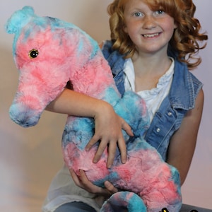May include: A young person is holding a large, plush seahorse toy. The seahorse is pink and blue with a sparkly gold eye. The toy is made of soft, fluffy fabric. The person is wearing a denim vest and jeans.