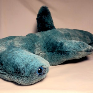 May include: A plush hammerhead shark toy in shades of teal and blue. The stuffed animal has a soft, furry texture, a distinctive hammer-shaped head, and a single blue eye. The toy is designed to resemble a hammerhead shark.
