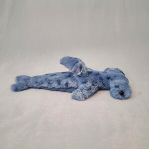 Pu&ograve; includere: Un giocattolo di peluche a forma di squalo martello blu con una superficie morbida e strutturata. Il giocattolo presenta una caratteristica testa a forma di martello, pinne e un piccolo occhio nero. Lo squalo &egrave; sdraiato su una superficie grigio chiaro.