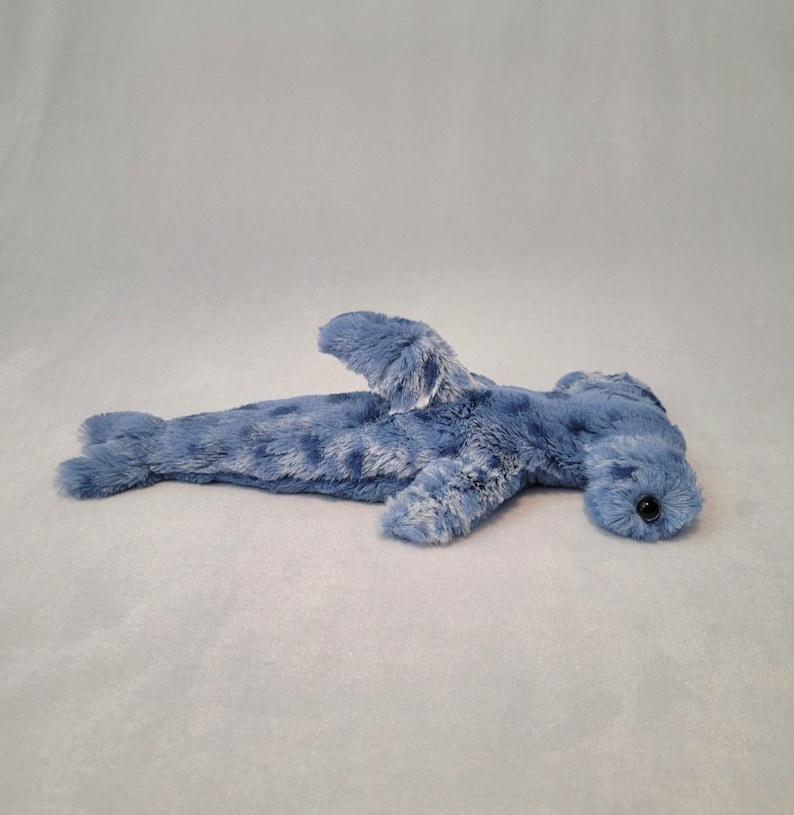 Pu&ograve; includere: Un giocattolo di peluche a forma di squalo martello blu con una superficie morbida e strutturata. Il giocattolo presenta una caratteristica testa a forma di martello, pinne e un piccolo occhio nero. Lo squalo &egrave; sdraiato su una superficie grigio chiaro.