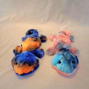 May include: Two plush toy salamanders in the image. One is blue and orange, the other is pink and blue. Both have black button eyes and are lying on a white surface. The toys are soft and fluffy.