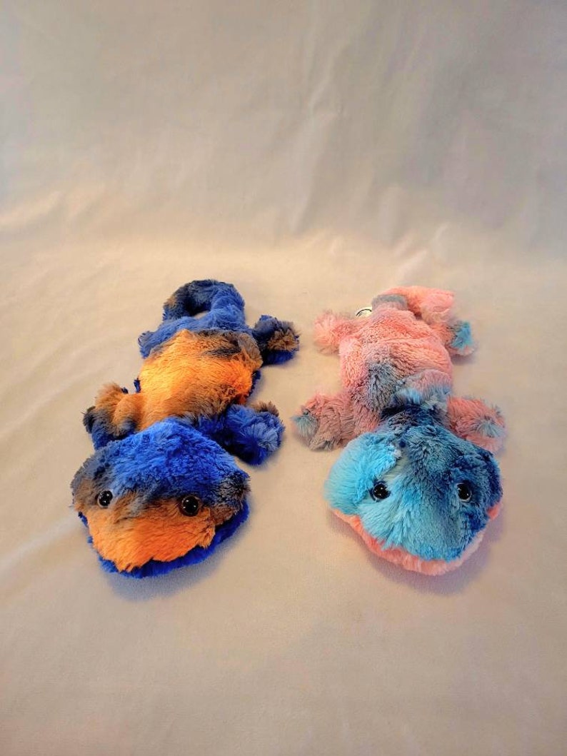 May include: Two plush toy salamanders in the image. One is blue and orange, the other is pink and blue. Both have black button eyes and are lying on a white surface. The toys are soft and fluffy.