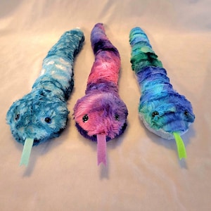 May include: Three plush snake toys in shades of blue, purple, and green. Each snake has a soft, textured body, black button eyes, and a contrasting colored tongue. The snakes are designed to resemble a snake.
