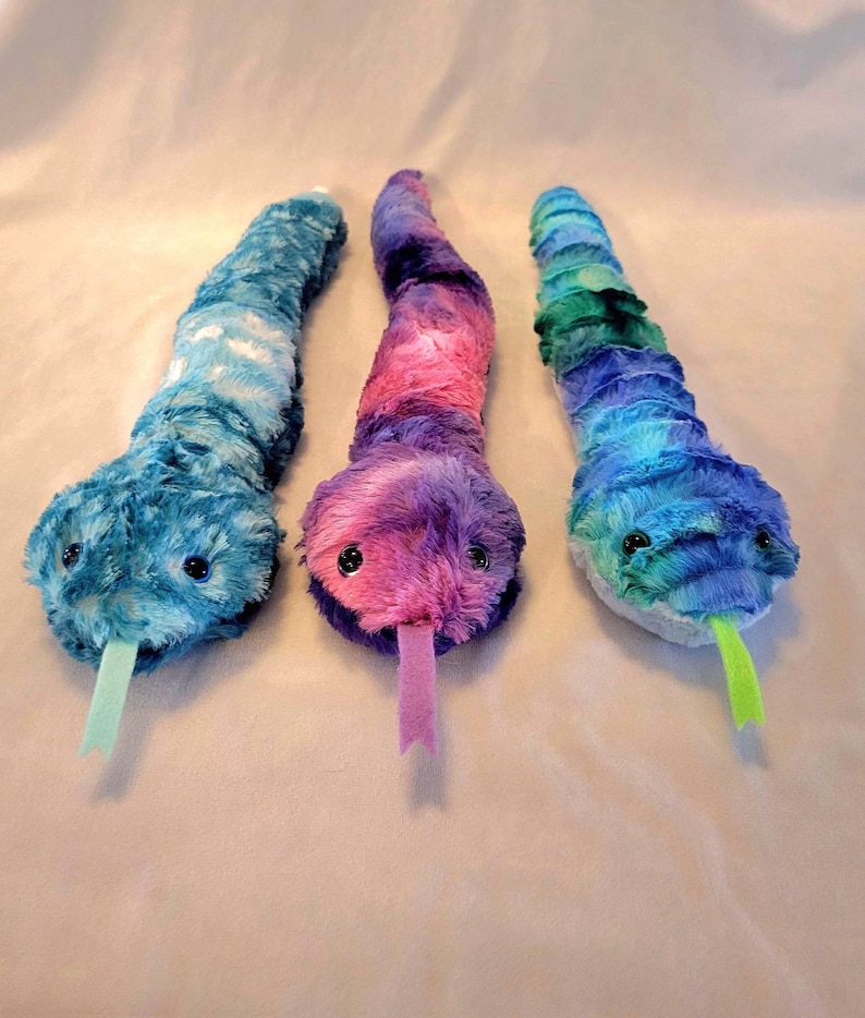 May include: Three plush snake toys in shades of blue, purple, and green. Each snake has a soft, textured body, black button eyes, and a contrasting colored tongue. The snakes are designed to resemble a snake.