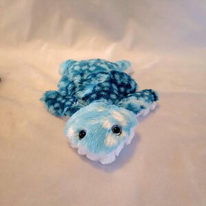 May include: A plush turtle toy in shades of blue and white. The turtle has a patterned shell and a light blue head with large, black eyes. The toy is soft and fluffy, designed for children.