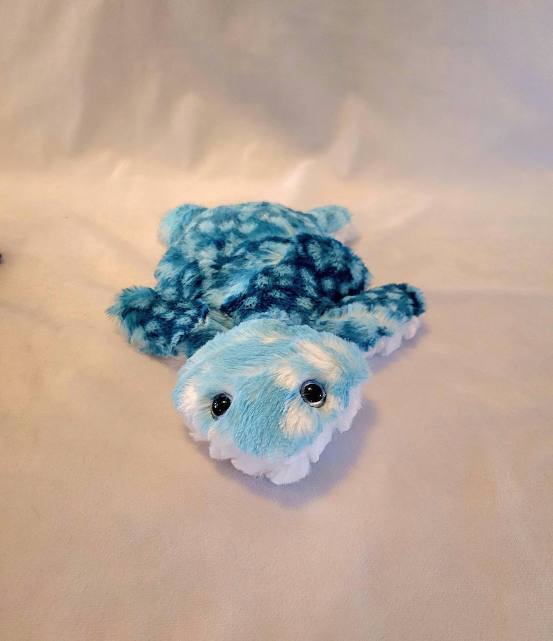 May include: A plush turtle toy in shades of blue and white. The turtle has a patterned shell and a light blue head with large, black eyes. The toy is soft and fluffy, designed for children.