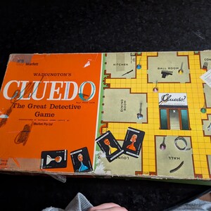 May include: A rectangular Waddington's Cluedo board game box featuring an orange left panel with white text and a yellow right panel depicting a floor plan of a mansion. The box includes illustrations of game cards, weapons, and room labels.