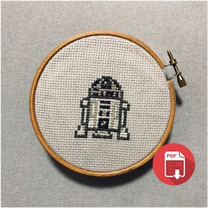 Könnte beinhalten: Ein Kreuzstich-Stickrahmen mit einem pixeligen Bild von R2-D2 aus Star Wars. Der Rahmen ist aus Holz und hat einen beigefarbenen Stoffhintergrund. Das Bild ist in Schwarz, Weiß und Rot.