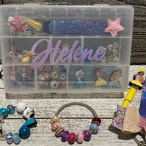 May include: A clear plastic organizer box with multiple compartments, filled with beads, charms, and accessories. The name "Helene" is written in purple script on the front. Various bracelets and keychains with charms are also visible.