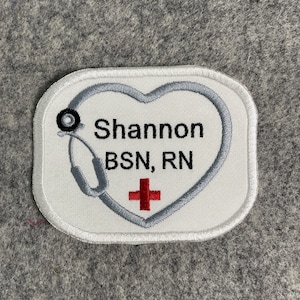 May include: Embroidered patch with a white background and a gray heart-shaped stethoscope design. The patch includes the name "Shannon" and the credentials "BSN, RN" in black text, along with a red cross symbol.