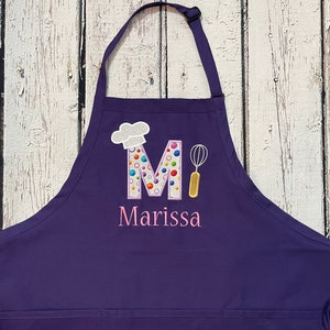 May include: Purple chef's apron with a white chef's hat, a colorful letter M, and a whisk. The apron is personalized with the name Marissa.