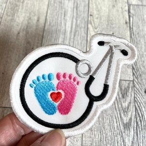May include: White embroidered patch with a stethoscope, two baby footprints, and a red heart. The footprints are blue and pink.