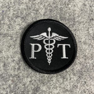 May include: A black circular patch with a white embroidered caduceus symbol and the letters "PT". The patch has a black border and is likely an emblem for physical therapy professionals.