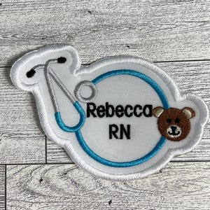 May include: White embroidered patch with a stethoscope, a teddy bear, and the text "Rebecca RN".