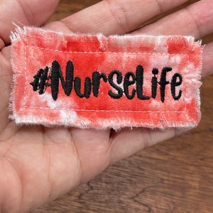 May include: A red and white tie-dye fabric patch with the text "#NurseLife" embroidered in black.