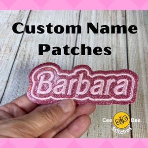 May include: Pink glitter iron-on patch with the name "Barbara" in white lettering. The patch is held in a hand. The text "Custom Name Patches" is at the top of the image. The text "Cee Bee Stitches" is at the bottom of the image.