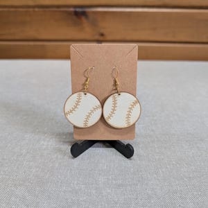 May include: A pair of round, flat wooden earrings featuring a white surface with a tan baseball stitch pattern. The earrings are attached to a rectangular brown card with gold-toned metal hooks, designed to mimic the appearance of a baseball.
